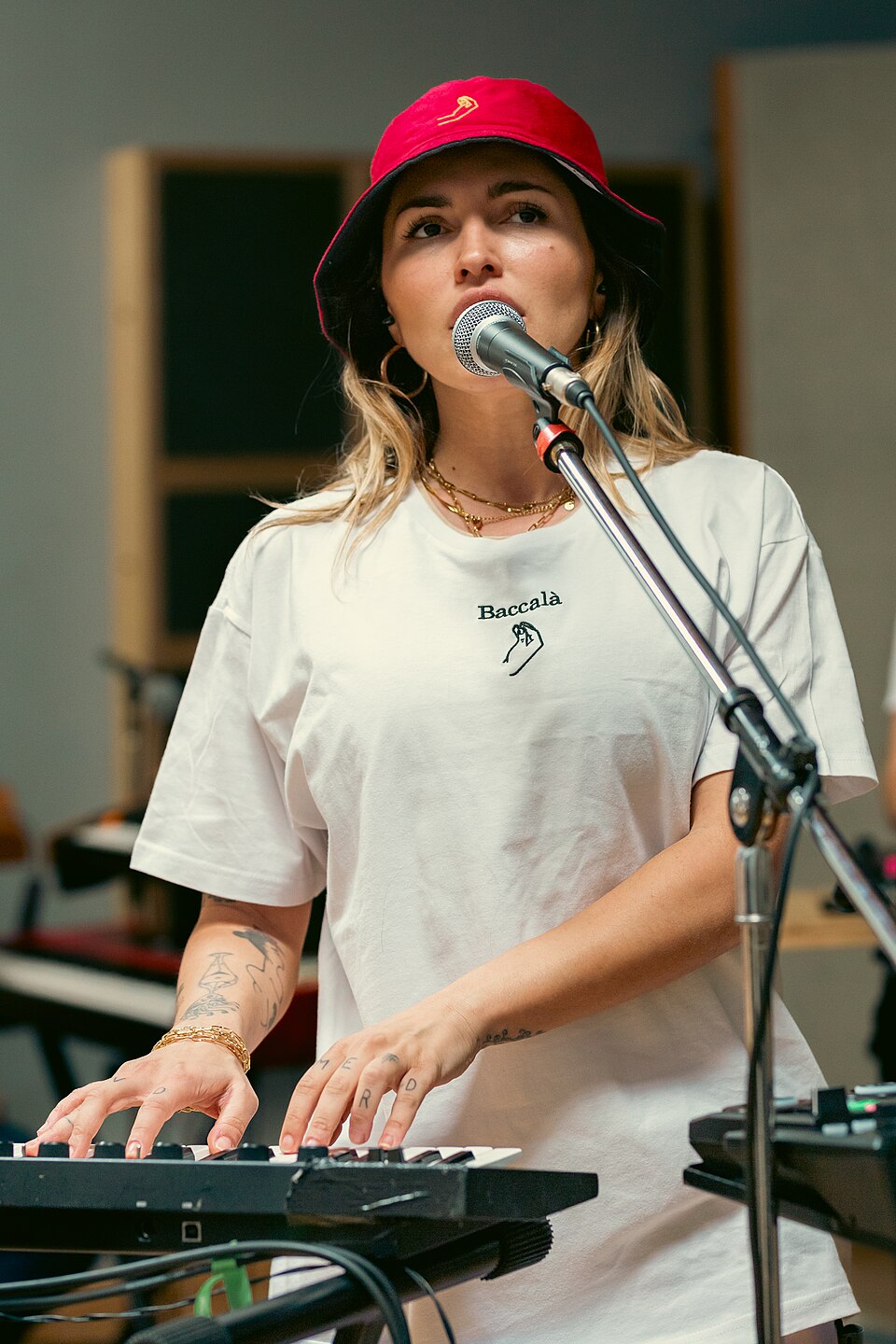Serena Brancale in studio, al microfono e alla tastiera, con cappello rosso e maglietta Baccalà; foto del 2024